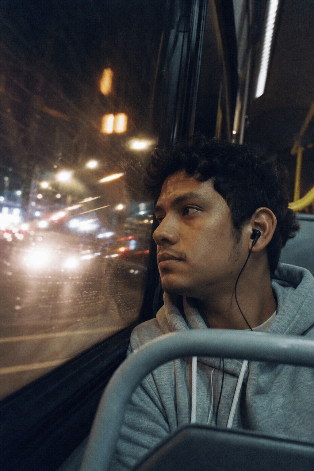 A young man rides a city bus at night, the lit street reflected in the window.