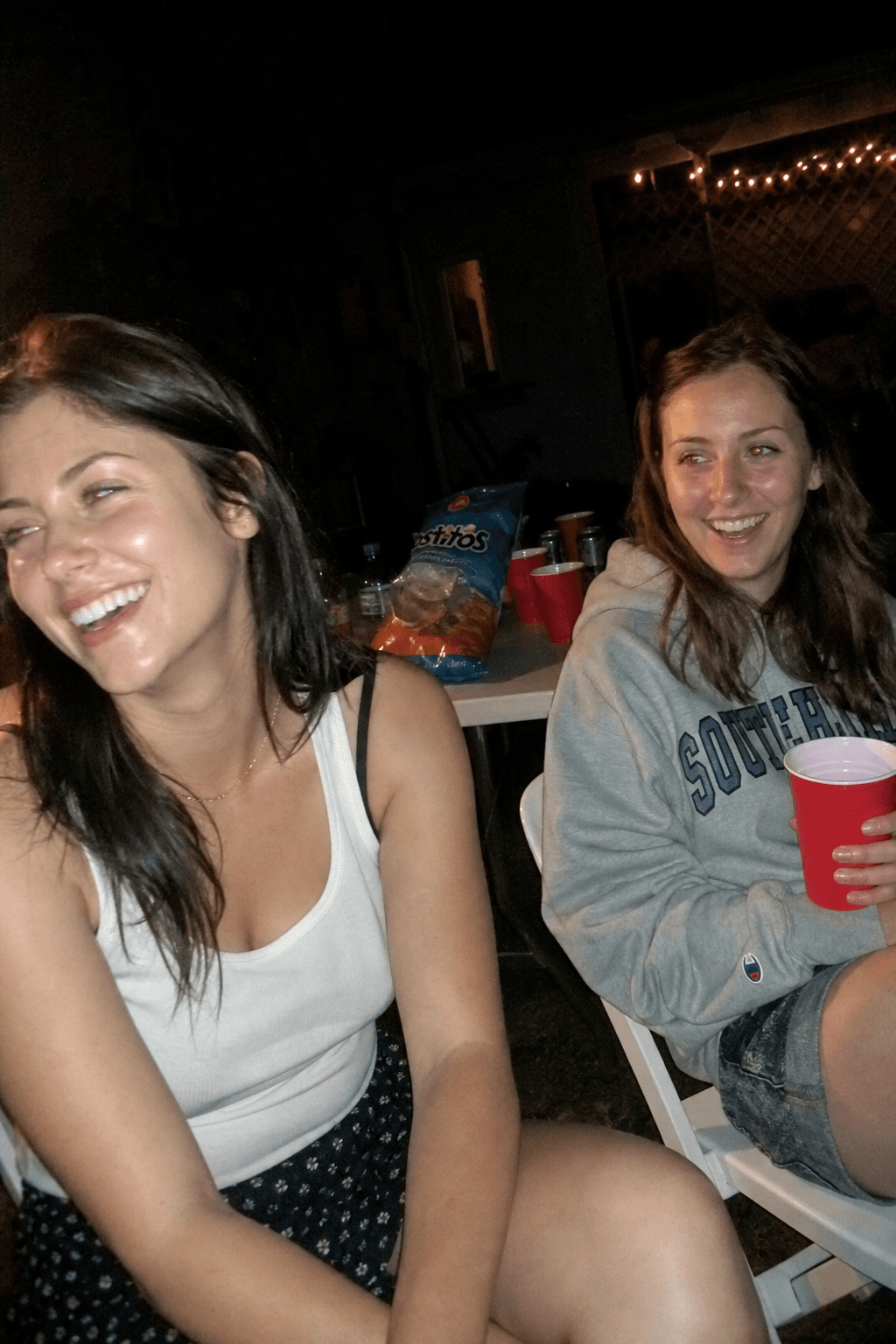 Two friends laugh at a backyard gathering, lit by a harsh on-camera flash.